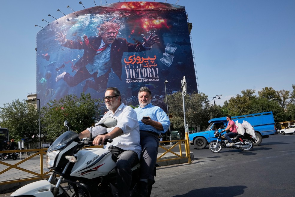 Motociclistas passam por um painel que retrata o presidente dos Estados Unidos, Donald Trump, debaixo d’água em meio a explosões, com os dizeres 'Grande vitória! Estreito de Ormuz', em Teerã, Irã, na segunda-feira, 24 de agosto de 2026 — Foto: AP Foto/Vahid Salemi