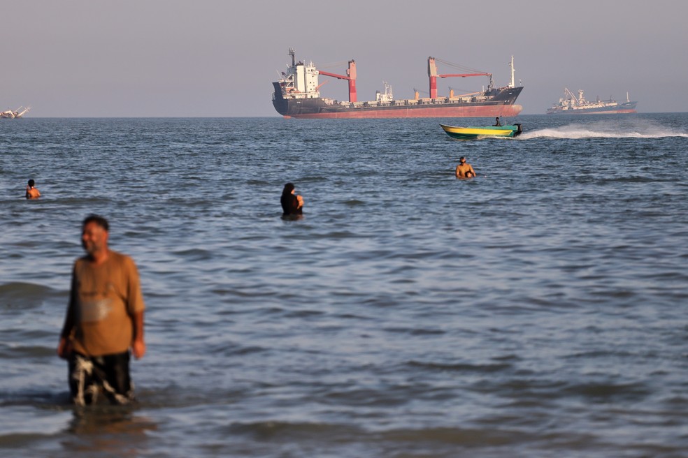 Moradores nadam nas águas do Estreito de Ormuz enquanto uma pequena lancha passa por navios de carga e outras embarcações comerciais perto de Bandar Abbas, Irã, na quarta-feira, 17 de junho de 2026 — Foto: Amirhosein Khorgooi/ISNA via AP