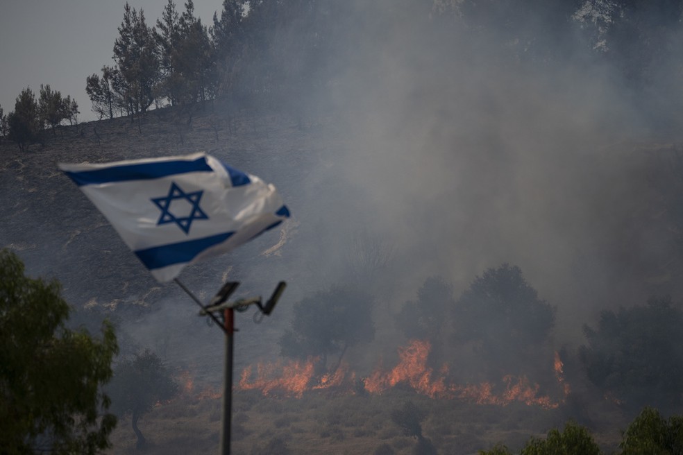 Bandeira israelense tremula ao lado de área incendiada, em Safed, norte de Israel e perto da fronteira com o Líbano, após ataque do Hezbollah, nesta quarta-feira, 12 de junho de 2024 — Foto: Leo Correa/AP